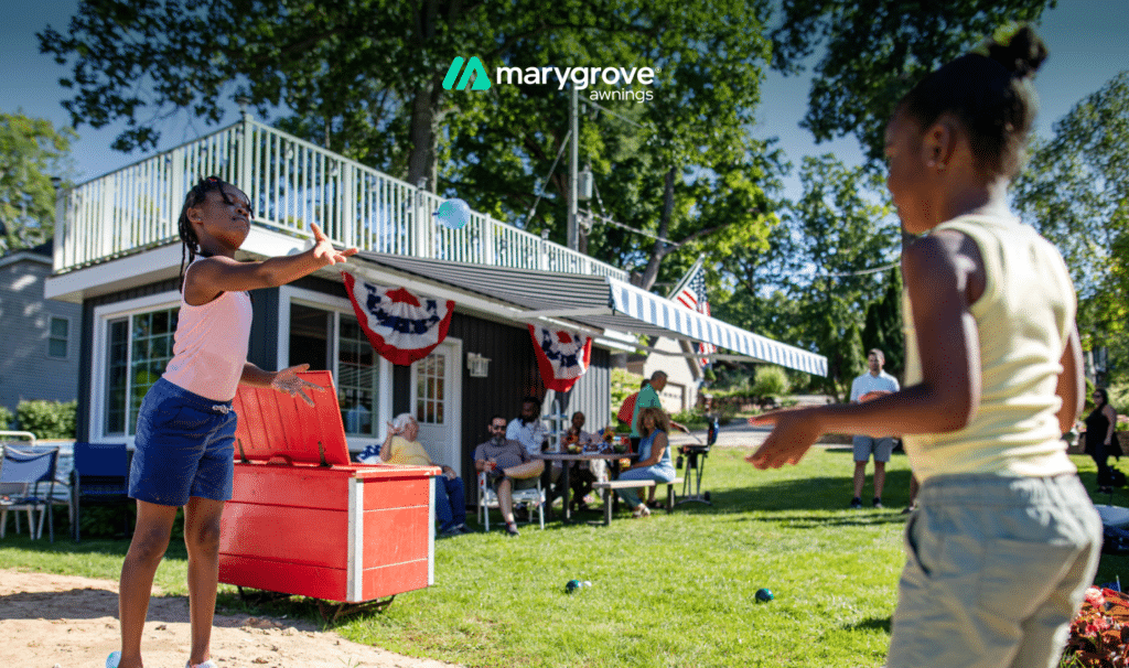 Two children play catch in a backyard while family members gather beneath a striped Marygrove awning.