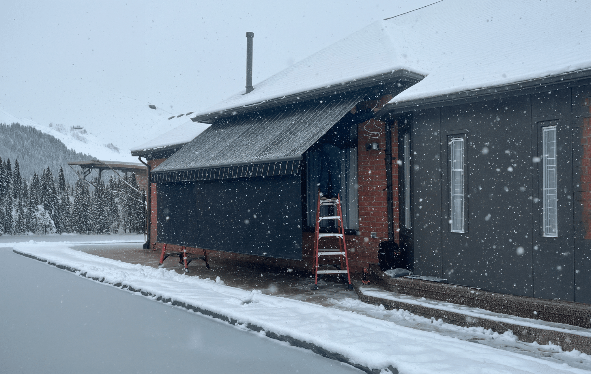Retractable awning extended over a building entrance during heavy snowfall, with snow-covered mountains in the background.